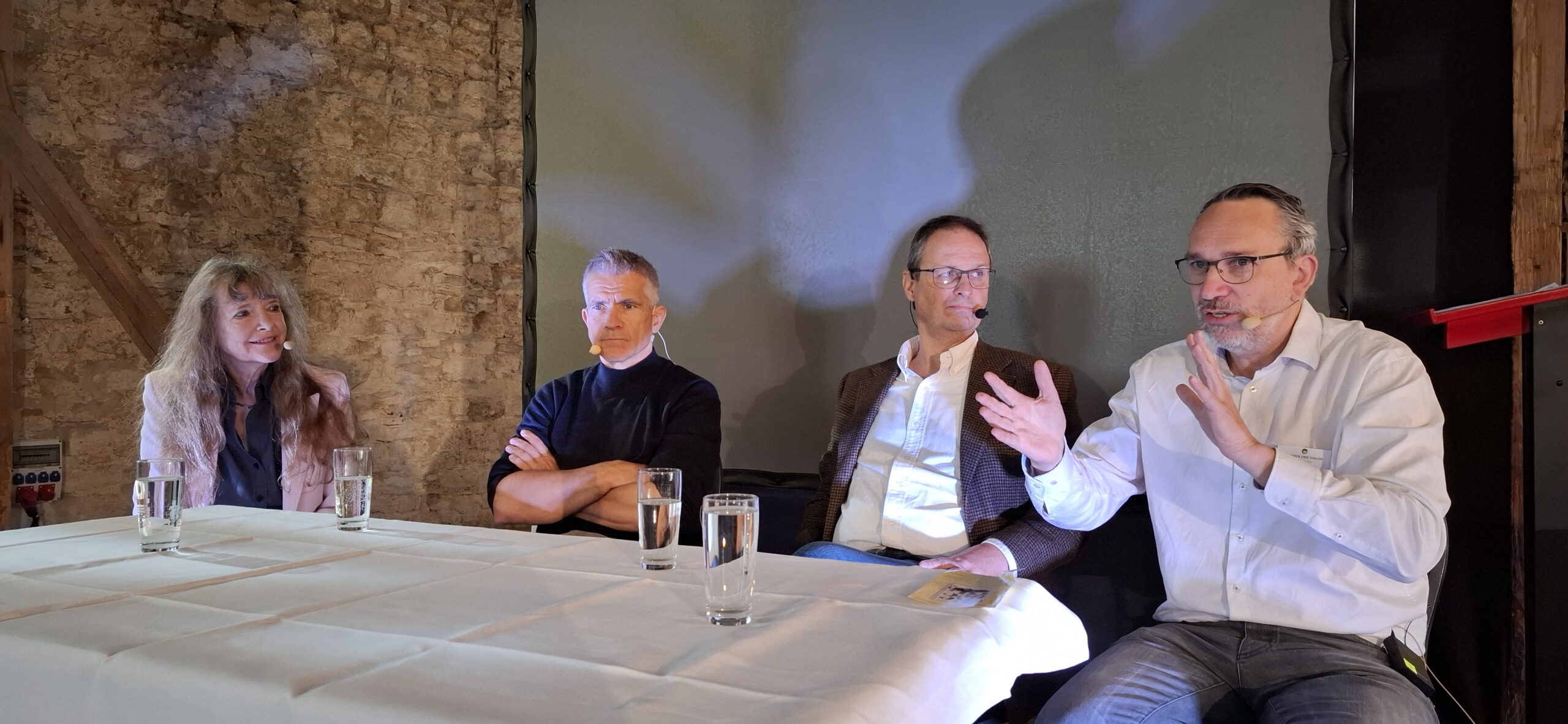 Vier Personen sitzen an einem mit weißem Tuch bedeckten Tisch, eine Person rechts gestikuliert während einer Diskussion.