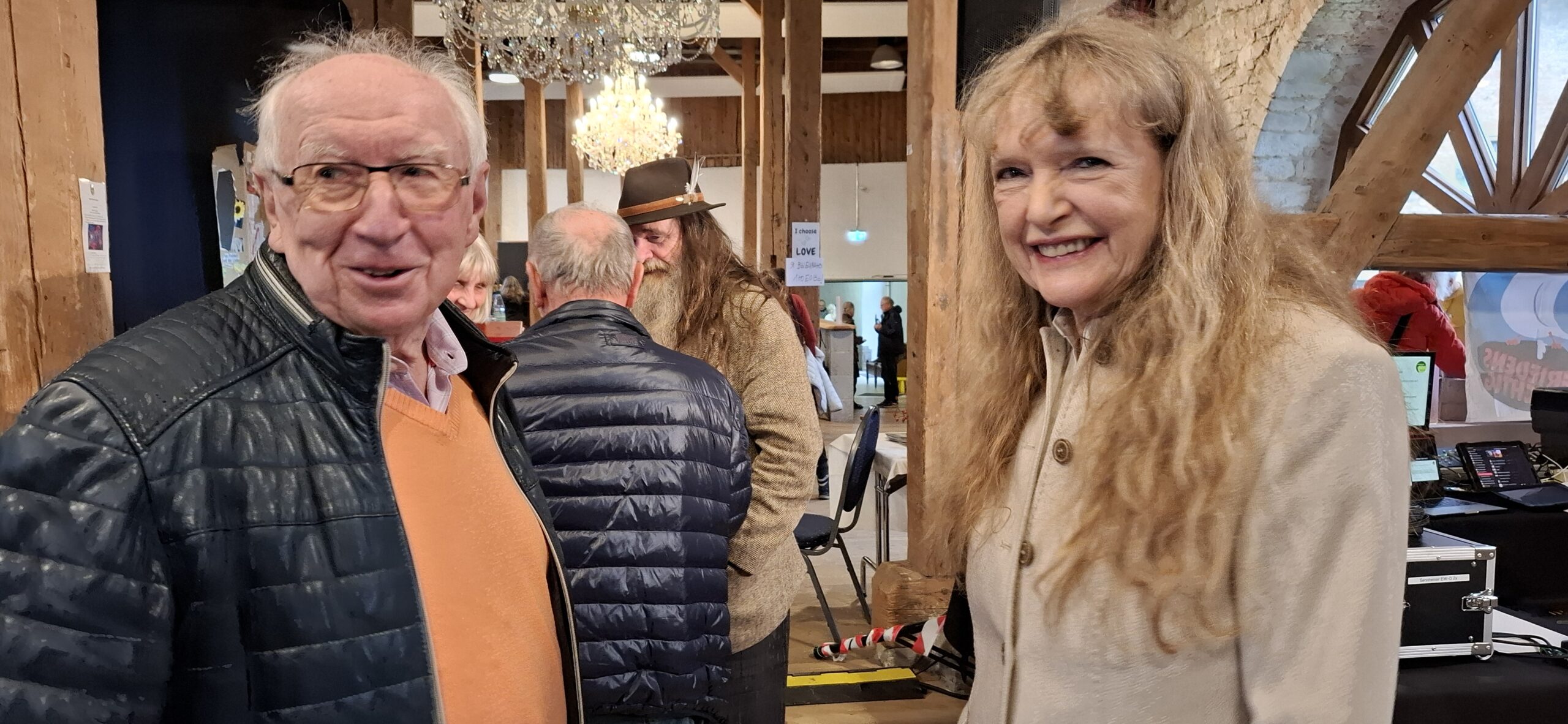 Zwei ältere Personen, ein Mann mit Brille und orangefarbenem Pullover unter schwarzer Jacke und eine Frau mit langen blonden Haaren und hellem Mantel, lächeln in einem Raum mit Holzbalken und Kronleuchter