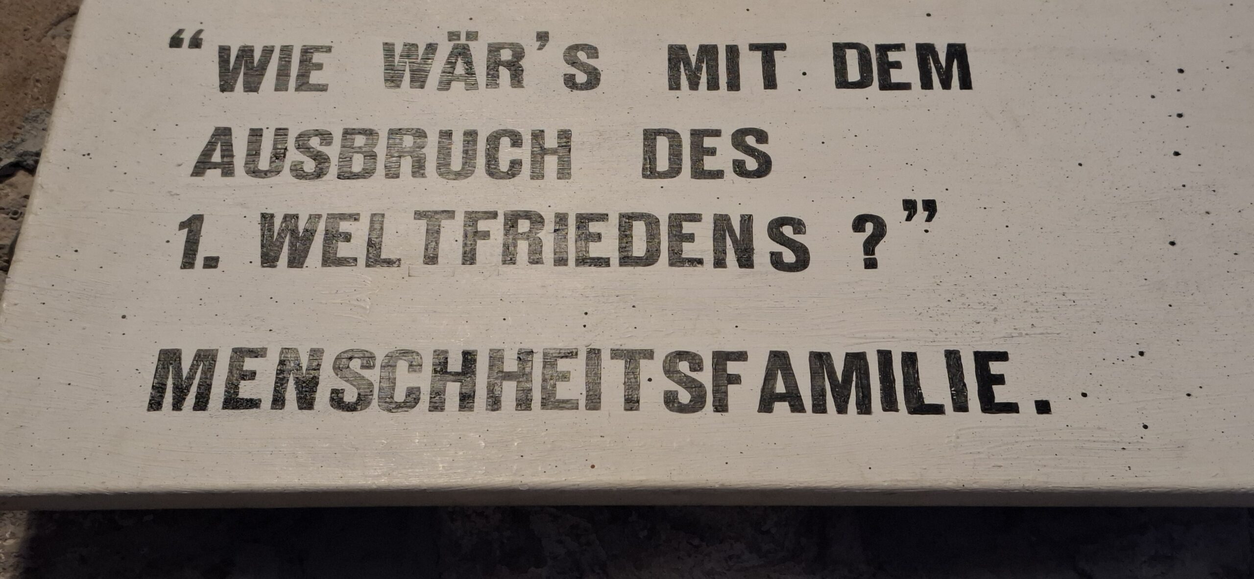 Schriftzug auf Betonplatte mit dem Text 'WIE WÄR'S MIT DEM AUSBRUCH DES 1. WELTFRIEDENS? MENSCHHEITSFAMILIE.'