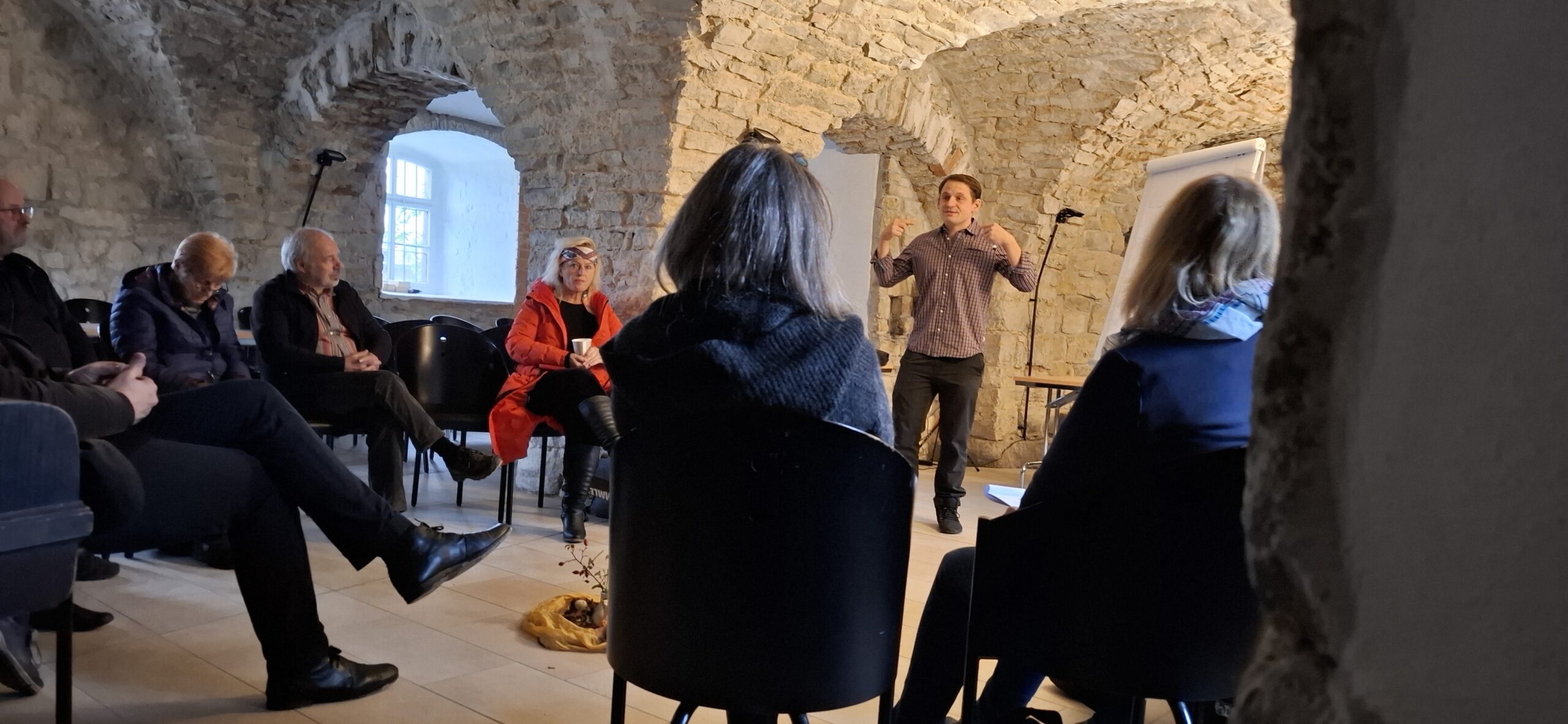 Mehrere Personen sitzen in einem historischen Gewölberaum im Halbkreis, eine Person steht und spricht vor einem Flipchart
